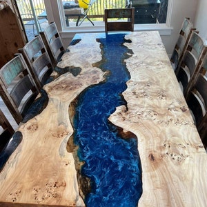 May include: A large wooden dining table with a blue epoxy river running through the center. The table is surrounded by rustic wooden chairs.