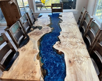 Pode incluir: Uma grande mesa de jantar de madeira com um rio de epóxi azul que atravessa o centro. A mesa é cercada por cadeiras de madeira rústicas.