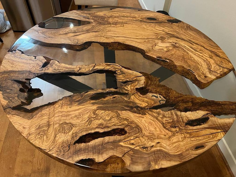 May include: A round wooden table top with a natural wood grain pattern and clear epoxy resin filling in the cracks. The table top is dark brown with light brown streaks and has a natural edge.