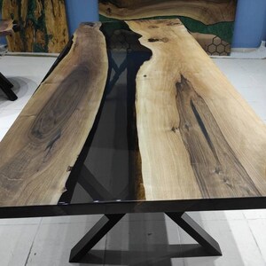 May include: A large wooden dining table with a black epoxy resin inlay. The table has a natural wood finish and a black metal X-shaped base.