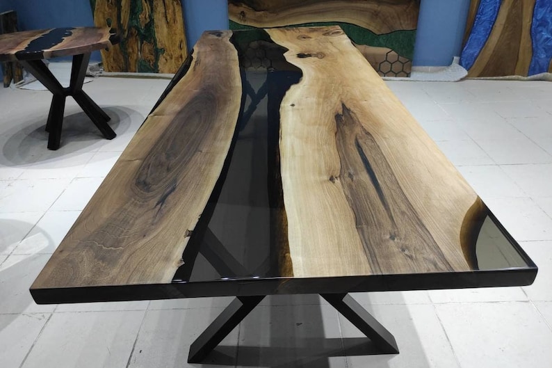 May include: A large wooden dining table with a black epoxy resin inlay. The table has a natural wood finish and a black metal X-shaped base.