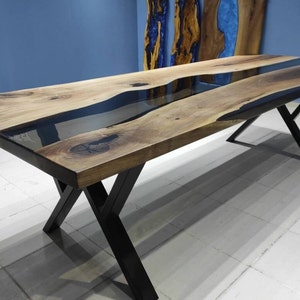 May include: A long wooden dining table with a black epoxy resin river running down the center. The table has a black metal X-shaped base.