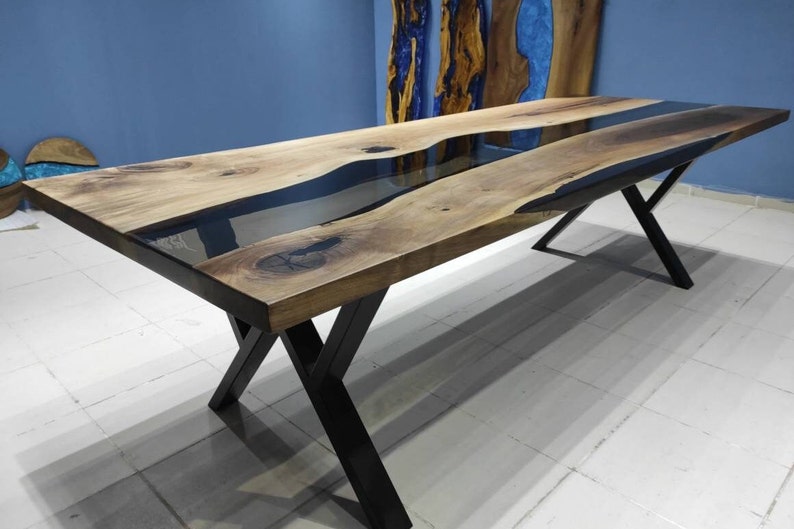 May include: A long wooden dining table with a black epoxy resin river running down the center. The table has a black metal X-shaped base.