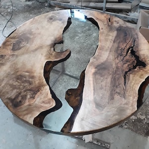 May include: A round wooden table top with a large, clear epoxy river running through the center. The wood is a light brown with dark brown streaks and knots. The epoxy is clear and smooth.