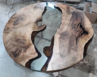 May include: A round wooden table top with a large, clear epoxy river running through the center. The wood is a light brown with dark brown streaks and knots. The epoxy is clear and smooth.