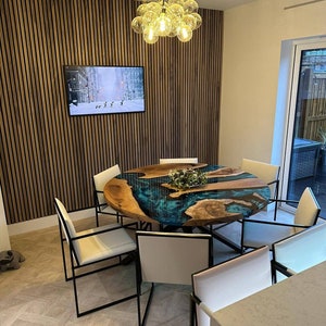 May include: A round dining table with a unique epoxy resin river table top in shades of blue and green. The table is surrounded by six white chairs with black metal frames. A modern chandelier with clear glass globes hangs above the table.