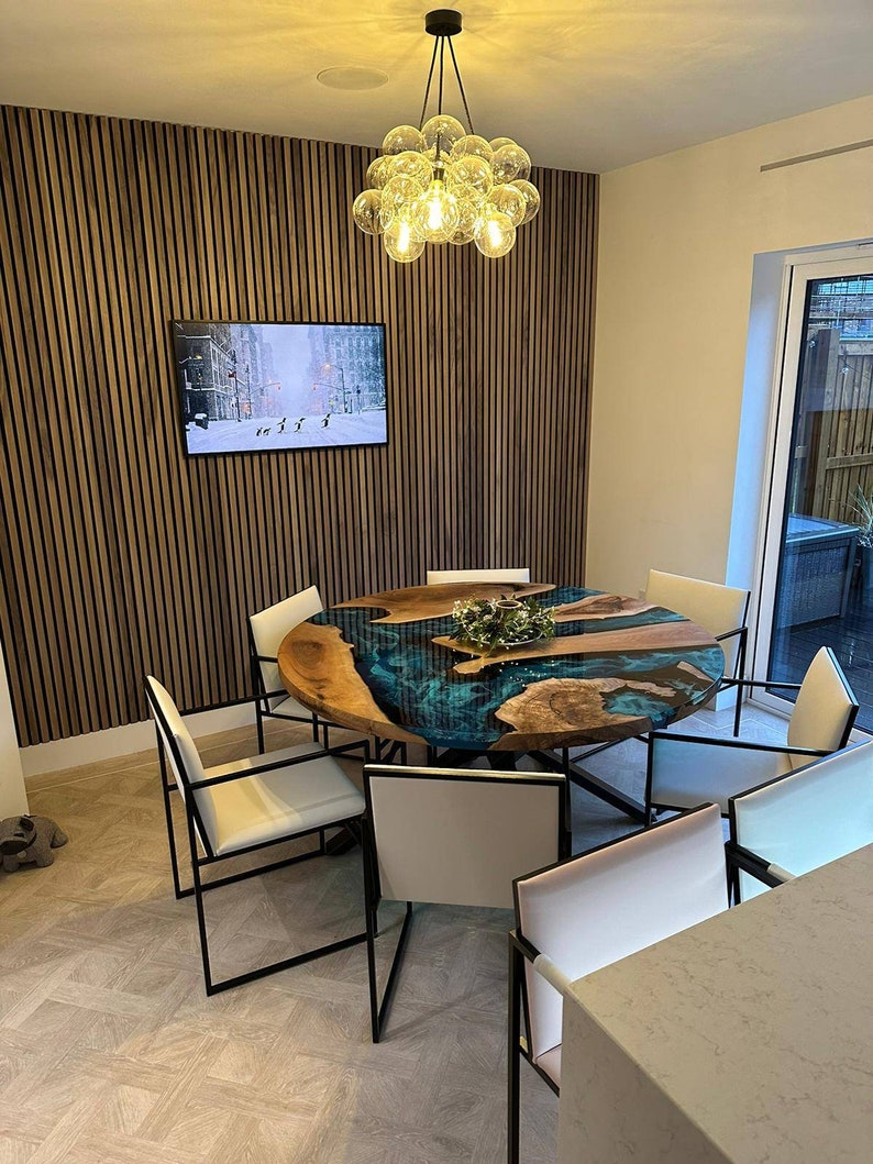 May include: A round dining table with a unique epoxy resin river table top in shades of blue and green. The table is surrounded by six white chairs with black metal frames. A modern chandelier with clear glass globes hangs above the table.