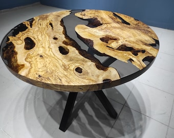 Könnte beinhalten: Ein runder Holztisch mit einzigartigem Design. Die Tischplatte hat eine hellbraune Holzmaserung mit dunkelbraunen Akzenten und einen klaren, dunkelgrauen Harzfluss. Der Tisch hat schwarze Beine.