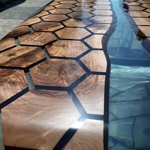Puede incluir: Una mesa de madera con un patr&oacute;n hexagonal negro incrustado en la madera. La mesa est&aacute; acabada con una resina epoxi transparente que crea una superficie lisa y brillante. Un r&iacute;o de resina azul fluye a trav&eacute;s del centro de la mesa.