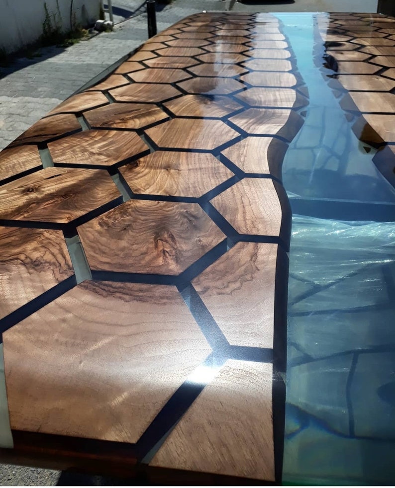 Puede incluir: Una mesa de madera con un patr&oacute;n hexagonal negro incrustado en la madera. La mesa est&aacute; acabada con una resina epoxi transparente que crea una superficie lisa y brillante. Un r&iacute;o de resina azul fluye a trav&eacute;s del centro de la mesa.