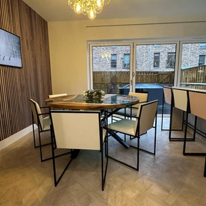 May include: A modern dining room with a wooden table and black metal chairs. The table has a green epoxy resin inlay and a natural wood finish. The chairs have white cushions and black metal frames. A chandelier with clear glass globes hangs from the ceiling.