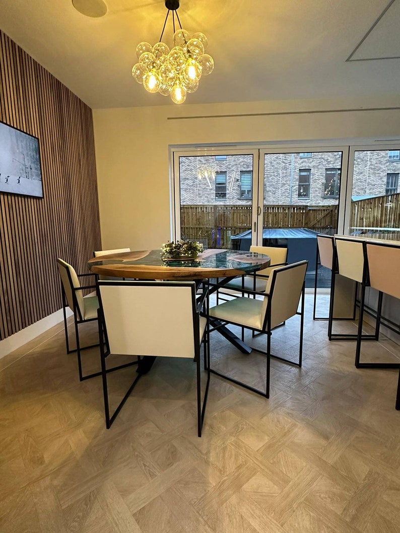 May include: A modern dining room with a wooden table and black metal chairs. The table has a green epoxy resin inlay and a natural wood finish. The chairs have white cushions and black metal frames. A chandelier with clear glass globes hangs from the ceiling.