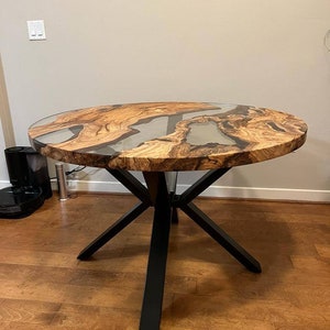 May include: Round wooden table with a clear epoxy resin top and a black metal base with four legs. The table has a natural wood grain pattern and the epoxy resin has a swirling pattern.