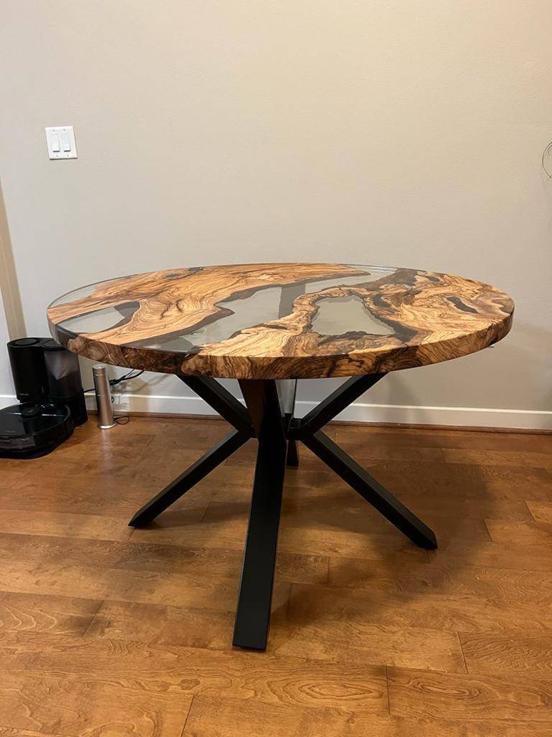 May include: Round wooden table with a clear epoxy resin top and a black metal base with four legs. The table has a natural wood grain pattern and the epoxy resin has a swirling pattern.