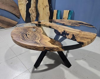 Könnte beinhalten: Ein runder Couchtisch mit einer Holzplatte und einem transparenten Harzeinsatz. Das Holz hat ein natürliches Maserungsmuster und wird von schwarzen Metallbeinen getragen. Zusätzliche Holzkunstwerke sind im Hintergrund zu sehen.