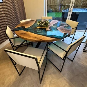 May include: A round wooden dining table with a blue and green epoxy resin inlay. The table has a black metal base and is surrounded by six white chairs with black metal frames.
