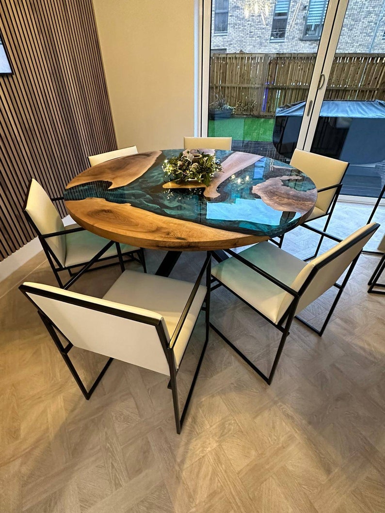May include: A round wooden dining table with a blue and green epoxy resin inlay. The table has a black metal base and is surrounded by six white chairs with black metal frames.