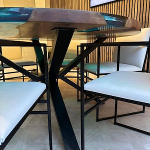 May include: A round wooden table with a clear epoxy resin top. The table has a black metal base with a unique X-shaped design. The table is surrounded by four white chairs with black metal frames.