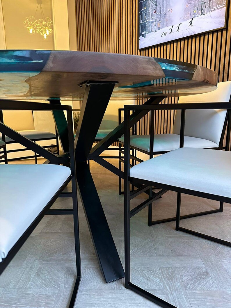 May include: A round wooden table with a clear epoxy resin top. The table has a black metal base with a unique X-shaped design. The table is surrounded by four white chairs with black metal frames.