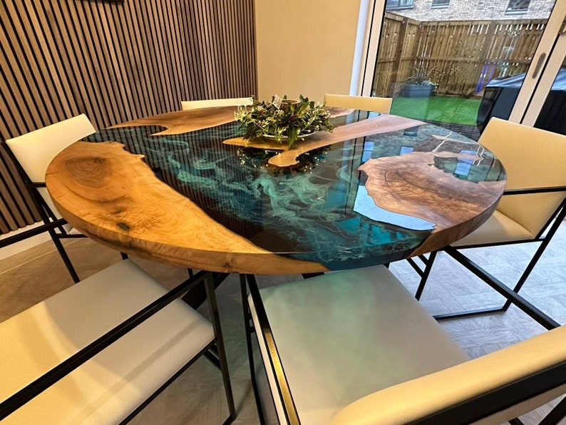 May include: A round wooden dining table with a teal blue epoxy resin inlay. The table has a natural wood finish and is surrounded by black metal chairs with white cushions.