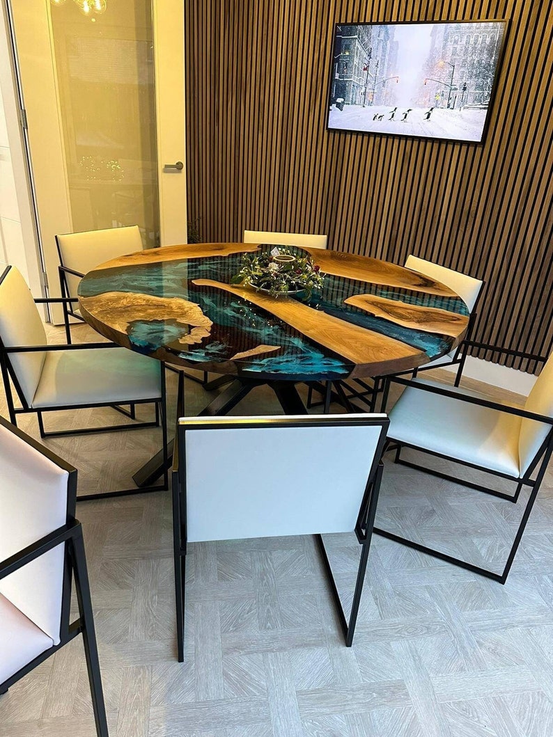 May include: A round wooden dining table with a blue and green epoxy resin inlay. The table has a black metal base and is surrounded by white chairs with black metal frames.