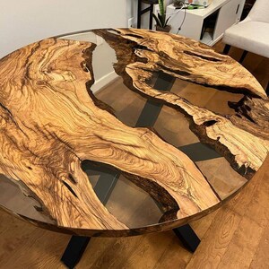 May include: A round wooden table with a clear epoxy resin inlay. The table has a black metal base with an X-shaped design. The table top has a natural wood grain pattern.
