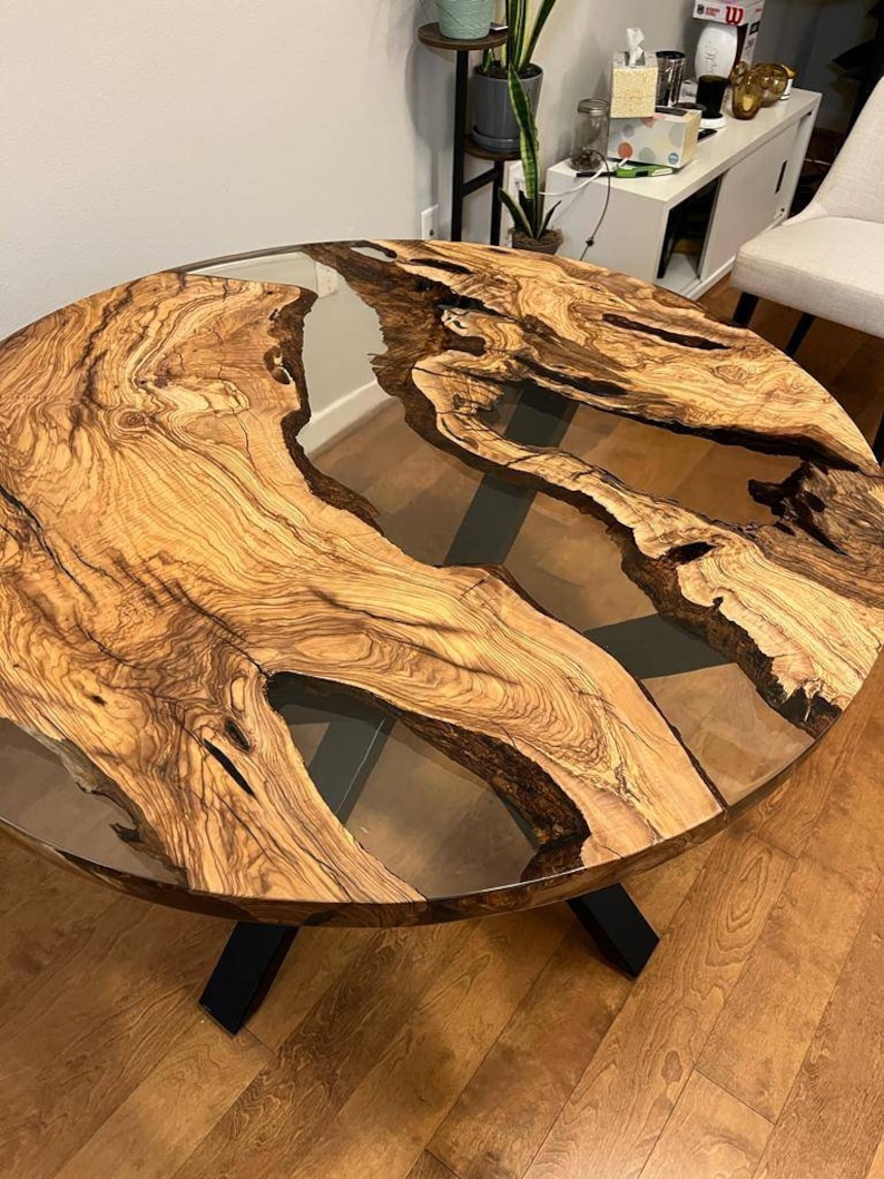 May include: A round wooden table with a clear epoxy resin inlay. The table has a black metal base with an X-shaped design. The table top has a natural wood grain pattern.