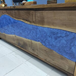 Puede incluir: Un armario de madera con tres puertas presenta una incrustación de resina azul que se asemeja a un río que fluye a través de la madera. El armario tiene un acabado de madera natural y está hecho de madera maciza.