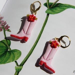 Könnte beinhalten: Ein Paar rosa Cowboy-Stiefel-Ohrringe mit roten Akzenten und goldenen Verschlüssen. Jeder Stiefel hat eine kleine rote Blume. Die Ohrringe sind vor dem Hintergrund eines Buches und einer grünen Pflanze platziert.