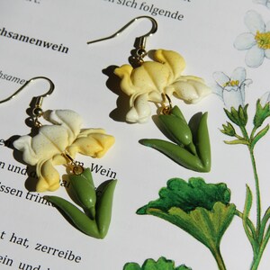 Puede incluir: Un par de pendientes hechos a mano con un diseño floral. Los pendientes tienen un gancho dorado y están adornados con colgantes en forma de flor crema y amarilla con detalles de hojas verdes. Los pendientes están sobre un libro con texto en alemán.