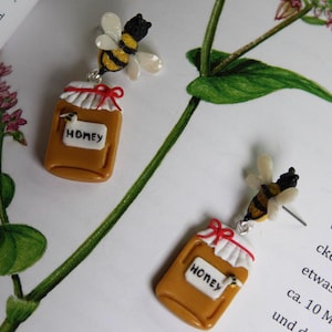 May include: A pair of novelty earrings featuring a honey jar and a bee. The earrings have a small, brown jar with a white lid and a red bow, labelled "HONEY". A bee with black and yellow stripes and white wings is attached.