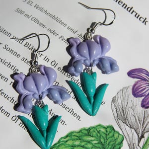 Puede incluir: Un par de pendientes de flores de iris lavanda con detalles de tulipanes verde turquesa. Los pendientes están suspendidos de ganchos plateados. Los pendientes están colocados sobre un fondo de texto e ilustraciones florales.