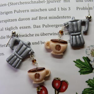 May include: Silver-colored espresso maker earrings with dangling cream-colored coffee cup charms. The earrings are on a page with text and floral illustrations. The coffee cup charms have a coffee bean design.
