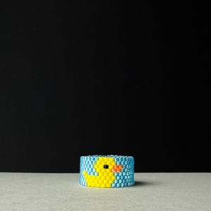 Könnte beinhalten: Ein Perlenring mit einem gelben Entenmuster auf blau-weißem Hintergrund. Der Ring ist zylindrisch und liegt auf einer hellgrauen Oberfläche vor einem schwarzen Hintergrund. Die Ente hat einen orangefarbenen Schnabel und schwarze Augen.