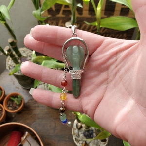 May include: A green angel pendant with a silver frame and a chain with seven chakra beads. The pendant is hanging from a hand.