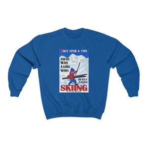 Pode incluir: Uma camisa de frio azul com uma ilustra&ccedil;&atilde;o vintage de uma menina esquiando. O texto diz "Era uma vez uma menina que realmente amava esquiar. Era eu. Fim."