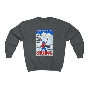 Pode incluir: Uma camisola cinzenta escura com uma ilustra&ccedil;&atilde;o vintage de uma menina a esquiar. A ilustra&ccedil;&atilde;o &eacute; num esquema de cores vermelho, azul e branco e apresenta o texto "Once Upon A Time There Was A Girl Who Really Loved Skiing It Was Me. The End."