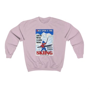 Pode incluir: Uma camisa de frio rosa claro com uma ilustra&ccedil;&atilde;o vintage de uma menina esquiando. O texto diz "Era uma vez, havia uma menina que realmente amava esquiar. Era eu. Fim."