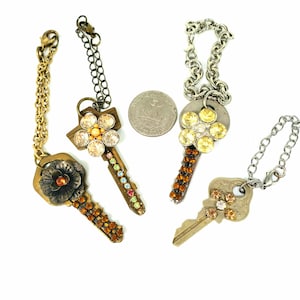 May include: Four decorative keychains with ornate designs. One features a flower with an orange center, another has a flower design with clear and orange gems, and two others have gem embellishments. Each keychain has a chain and clasp.