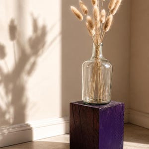 May include: A clear glass bottle containing a bouquet of dried beige bunny tail grass stems, positioned atop a solid, dark purple wooden block with a visible grain texture and a square, box-like shape.
