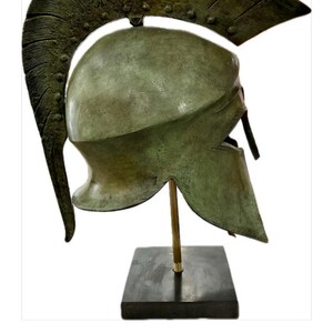May include: A bronze-colored Spartan helmet with a large, curved crest. The helmet is mounted on a black square base with a gold-colored rod. The helmet's design includes a face guard and a rounded top.