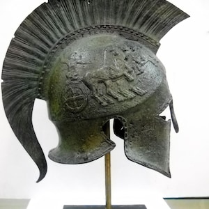 May include: A bronze Greek helmet with a crested plume and a relief of a chariot scene. The helmet is on a black stand.