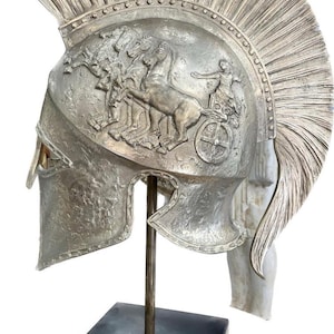May include: A silver-colored Spartan helmet with a detailed relief depicting a chariot race. The helmet features a prominent crest and is displayed on a black stand. The helmet's design is reminiscent of ancient Greek artistry.