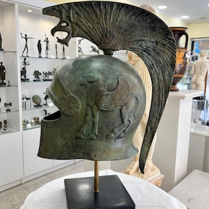 May include: Bronze-colored helmet with a winged horse design and a decorative crest featuring a stylized eagle head. The helmet is mounted on a black square base with a brass rod. The helmet is a historical or artistic item.