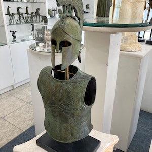 May include: Bronze sculpture of a Greek warrior bust wearing a helmet and armor. The sculpture is on a black marble base.