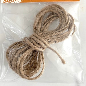 May include: A package of jute twine for hanging. The twine is a natural brown color and is coiled inside a clear plastic bag. The packaging includes a brown and white label with the text "JUTE TWINE" and the company name.