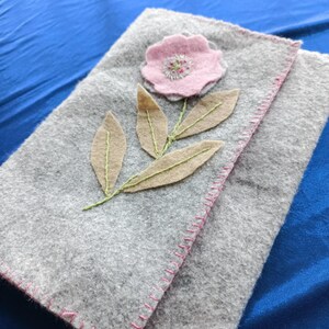 May include: A rectangular grey felt pouch featuring a pink felt flower appliqué with embroidered details and light brown felt leaves. The edges of the pouch are finished with a decorative pink blanket stitch, creating a textured and handcrafted appearance.
