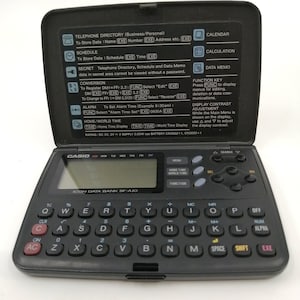 Pode incluir: Uma calculadora preta Casio Icon Data Bank SP-A10 com calendário, relógio e armazenamento de dados integrados. A calculadora tem um teclado QWERTY e uma pequena tela LCD.