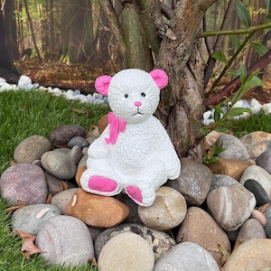 May include: A white teddy bear ornament with pink ears, paws, and a bow. The bear is positioned amongst rocks and grass, with a tree and a forest backdrop. The figurine has a textured surface.
