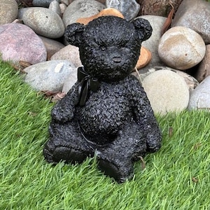 May include: A black teddy bear statue with a textured finish and a black bow. The bear is positioned on green grass, with rocks of different sizes in the background. Suitable for garden or home decor.